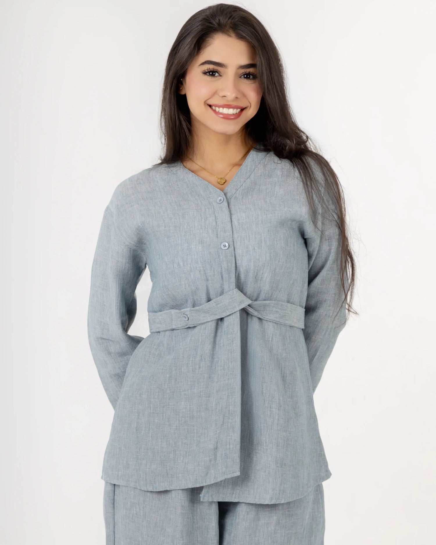 A woman modeling a light gray linen two-piece set consisting of a long-sleeve belted top and matching wide-leg trousers, photographed against a plain white studio background.